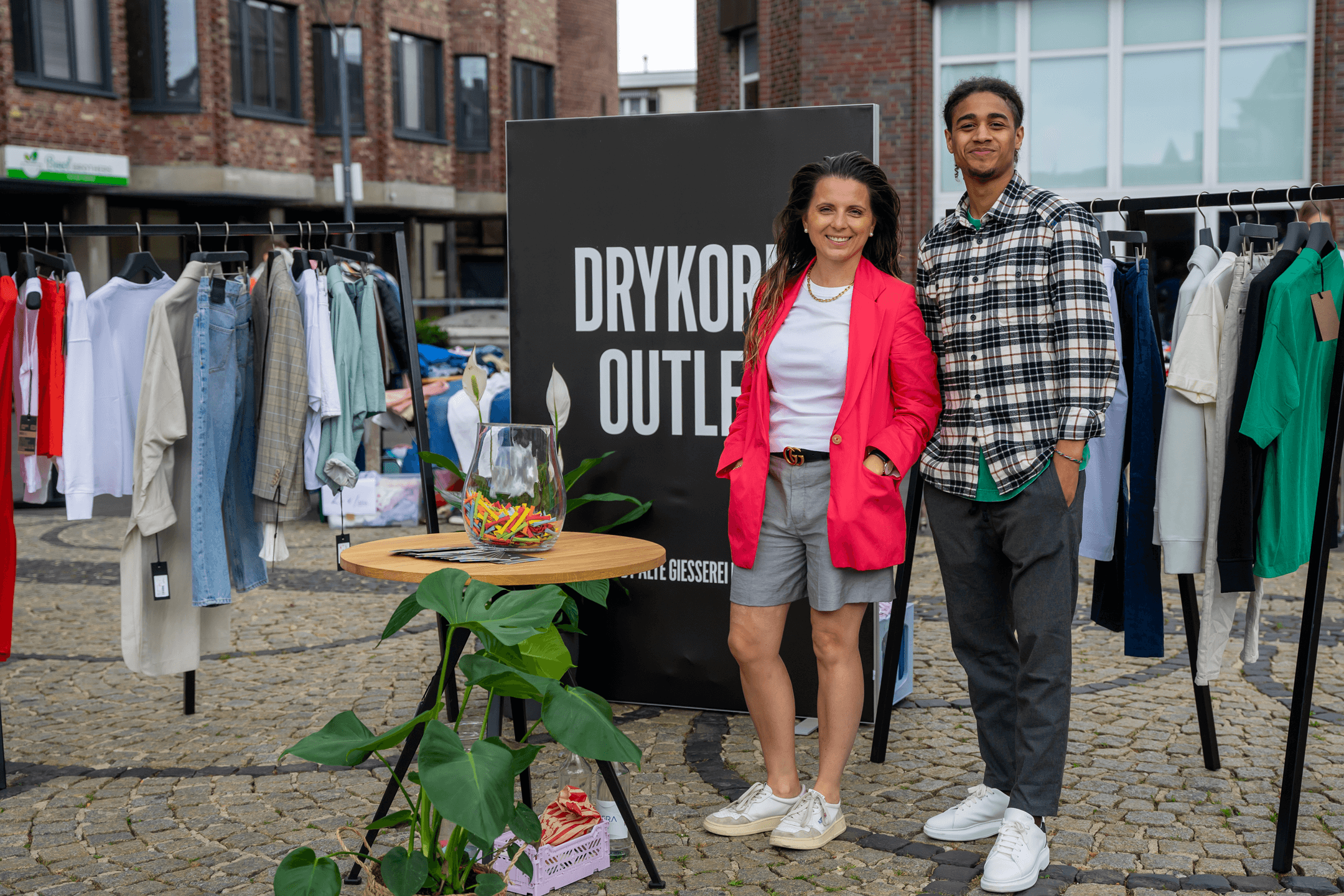 Zwei Personen, eine Frau und ein Mann, stehen lächelnd nebeneinander vor einem großen schwarzen Schild mit der Aufschrift