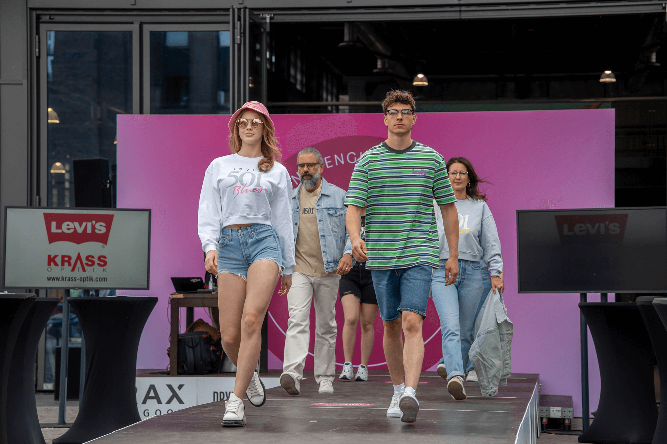 Mehrere Models laufen auf einem Laufsteg im Freien, während des 2. Mönchengladbacher Fashion Day, bei einer Modenschau. Im Vordergrund sind eine junge Frau und ein junger Mann zu sehen. Die Frau trägt einen rosa Sonnenhut, eine Sonnenbrille, ein weißes Sweatshirt mit dem Aufdruck