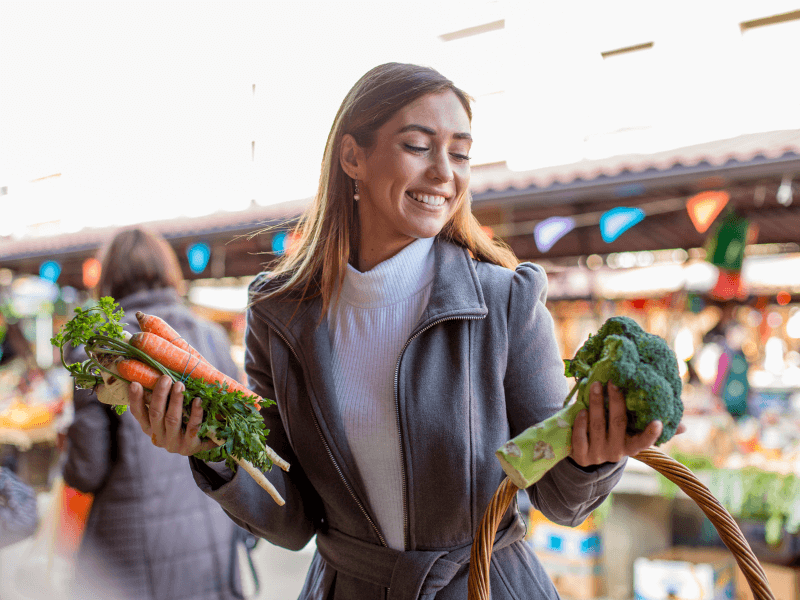 Eine junge Frau die lächelnd auf einem Wochenmarkt steht, bekleidet mit einem weißen Pullover und einem grauen Mantel. In ihrer linken Hand hält sie frisches Staudengemüse, bestehend aus Möhren und Rüben, in ihrer rechten Hand hält sie frischen Brokkoli und den Henkel eines Einkaufkorbs.