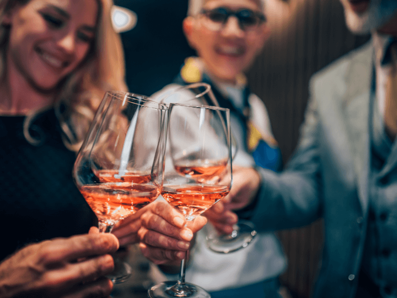 Im Vordergrund des Bildes sieht man mehrere Weingläser, gefüllt mit einem Roséwein, die von einer Gruppe von Menschen zum Anstoßen zueinander geführt werden. 