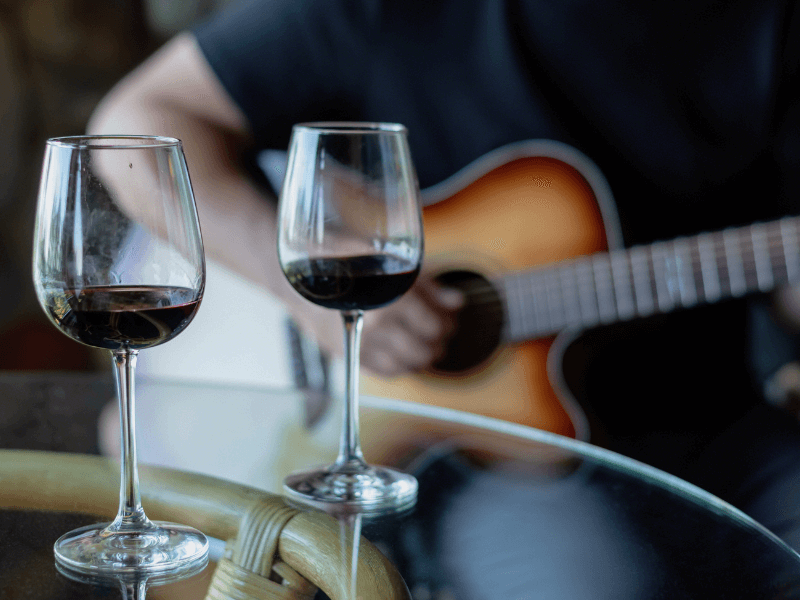 Nahaufnahme von zwei Weingläsern, gefüllt mit Rotwein, die auf einem runden Glastisch stehen, im Hintergrund sieht man einen Teilausschnitt einer Gitarre, die von einem Mann gespielt wird.