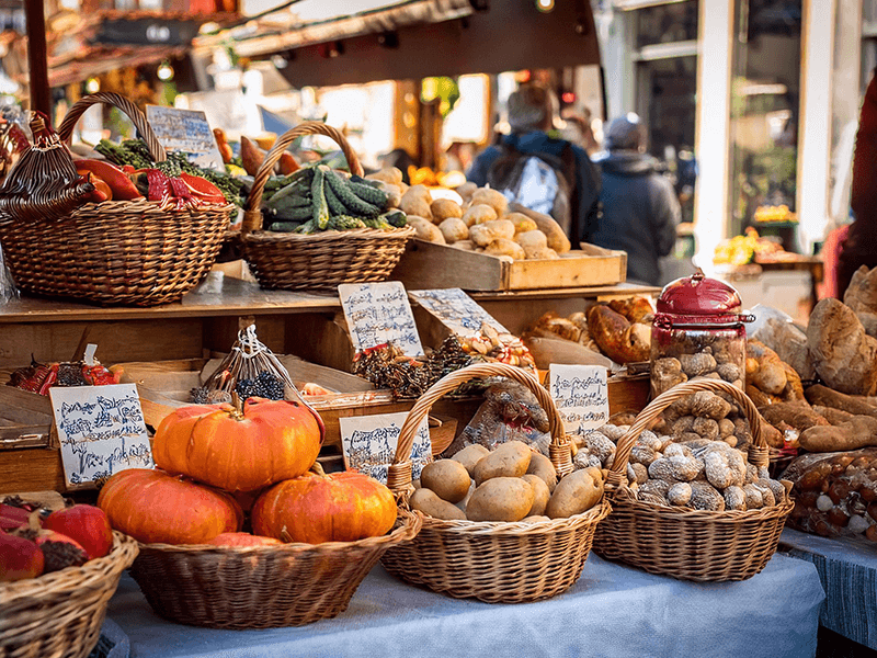 Bunter Marktstand mit Körben voller frischem Gemüse und Früchten wie Kürbissen, Kartoffeln, Paprika und Gurken sowie verschiedenen Backwaren und verpackten Lebensmitteln. Im Hintergrund sind verschwommene Umrisse von Personen und weitere Marktstände zu sehen.